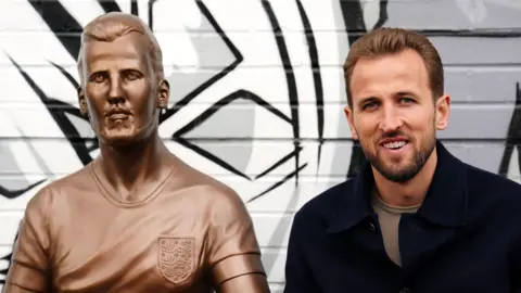 PA Media Harry Kane is seen sitting to the right of his statue. Both are seen from the chest up. The brown statue depicts him wearing an England shirt with a neutral expression while the real Harry is smiling.