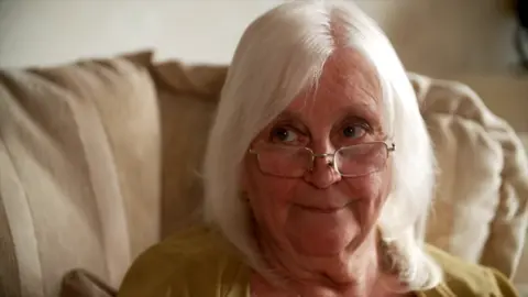 BBC Sue is sitting on her cream-coloured sofa. She has white shoulder length hair and a pair of glasses perched on her nose. She is wearing a mustard top.