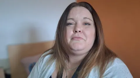 BBC Charmaine Denton looking into the camera, smiling slightly. She has long straight brown/blond hair and is wearing a white cardigan and a silver chain. She is sat on the bed in her bedroom in the shared recovery focused residence she lives in.