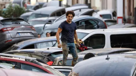 EPA A man stands on a pile of cars swept down a road by flood waters