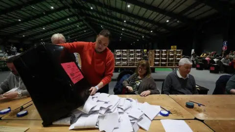 PA Media A woman wearing a red top empties a large black bin containing ballot papers onto a desk. To her right, a woman and a man both sit at the desk ready to count. They are in a large hall. In the background a number of other people sat at desks can be seen.