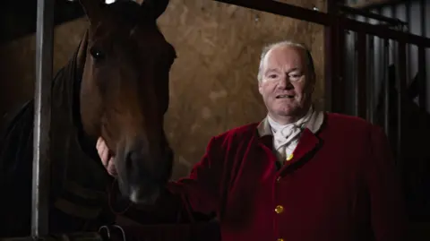 BBC A huntsman in red jacket looks at the camera with his horse