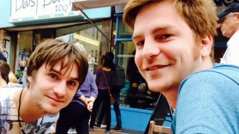 Simon Byrne Two young men smile in a busy street outside some shops. 