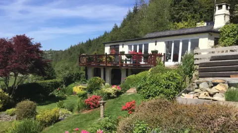 Aled Evans A view of the house, with a tiered garden. It is a white building on one level with a mahogany coloured balcony and a forest behind it  