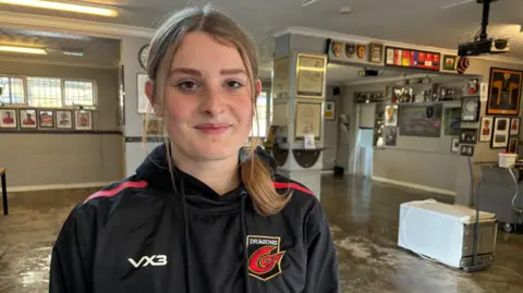 BBC A young lady with blonde hair tied back in a ponytail is looking at the camera. She's wearing a black hoodie with the Dragons rugby team logo on it. In the background is room that has been affected by floodwater. There's a white fridge on the floor to the right of frame and there are many different types of photo frames and shields on the walls. 