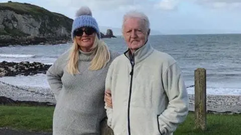 Handout A woman in a blue hat and grey sweater linking arms with a man with grey hair in a cream zip up. They are standing in front of a pebbled beach with rocks in the background. 