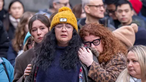 PA Media An emotional Ashley Storrie watching the hearse carrying her mum pass through Edinburgh. She is wearing a yellow hat and glasses and is being held by two other women, one of whom is red-headed and crying.