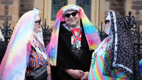 PA Media Three mourners dressed as colourful nuns, wearing rainbow habits and sunglasses and rainbow lanyards around their necks