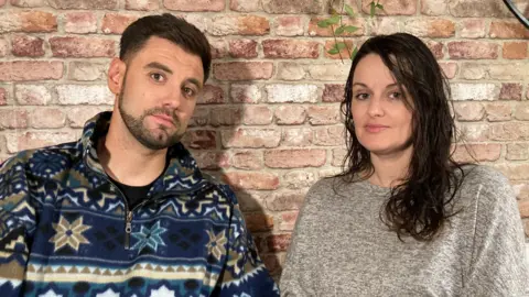 BBC/Carla Fowler A dark-haired man, wearing a colourful fleece, and a dark-haired woman, wearing a grey top, stand against an indoor brick wall