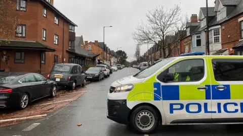 BBC A police van is parked at the end of a road of terraced houses with cars parked either side