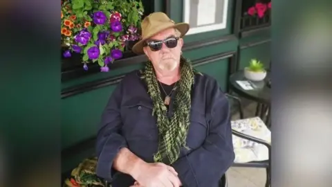 Police handout A man sitting outside. He has his hands crossed in front of him and his looking at the camera. He is wearing a hat, sunglasses, scarf, necklace and blue jacket. Behind him there is a table and chair and a building with a hanging basket of flowers on its wall.