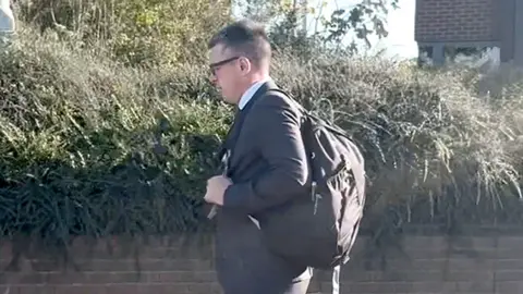 PA Media Christopher Bell has short black hair. He is wearing a black suit and black glasses and is walking on a street. He is also wearing a black backpack.