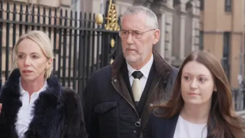 BBC Ian Harvey, with short grey hair and a grey beard and wearing a dark blue coat, is pictured walking to Liverpool Town Hall flanked by two women, one blonde and wearing a fur style coat and one with long brown hair and wearing a dark blue blazer.