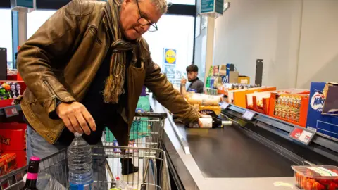 Alamy A middle aged man wearing a brown leather jacket and striped scarf loading a trolley of shopping on to the conveyor belt at Lidl