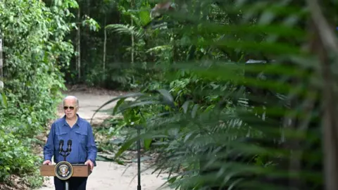 AFP Joe Biden makes statements to the news media from a podium in the Amazon rainforest. He is surrounded by greenery and tropical trees and brush.  