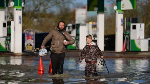 PA Media A man and a young boy carrying fishing rods wade through knee-high floodwater near petrol station pumps 