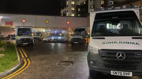 BBC Ambulances with green and yellow checked markings, with lights on, parked outside a hospital building in the dark.