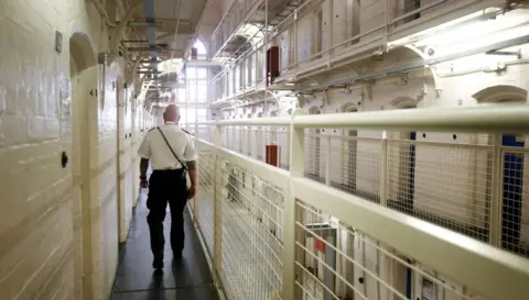 PA Media A prison officer walks away from the camera down the hallway of a prison. It's an open hallway so you can look up towards other floors. The walls are white and the fencing on the other side of the walkway towards the middle is also painted white. 