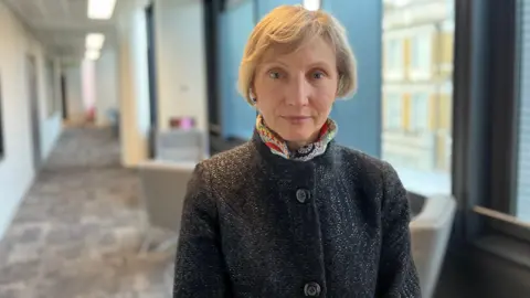 BBC Marina Litvinenko. She has short blonde hair and is wearing a dark grey coat with a colourful, patterned scarf around her neck. She is looking directly at the camera and smiling. 