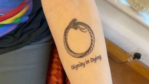 Mark Ansell/BBC A woman's forearm with a tattoo of a snake. Underneath the image are the words "dignity in dying". The woman is wearing a black T-shirt with a rainbow printed on it and blue trousers.