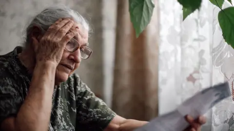 Getty Images Older woman sits in front of curtains looking at paperwork.