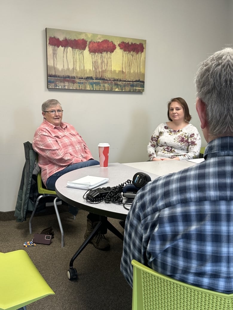 An adult woman with white hair sits at a round table speaking with an adult woman with brown hair, and a man with short white hair. The man is seen only from the back.