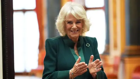 Getty Images Queen Camilla during a reception for winners of the Queen's Commonwealth Essay Competition at Buckingham Palace. She is wearing green, with a green brooch on her left lapel. She appears to be mid-clap and is smiling