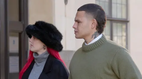 PA/Joe Giddens Tyron Frampton (right), known as Slowthai, walking outside a court wearing a green jumper. He has a tattoo on his neck and a short mullet haircut. A woman with long red hair and a furry hat walks beside him.