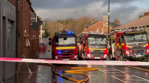 BBC Fire appliances parked three abreast in Kings Row, Coalisland, on Saturday. The red-brick street beyond is flooded with water reaching just below the rear number plate of red car.  Hoses are lying on the ground and the street is taped off. 