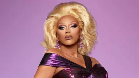 BBC/World of Wonder/Nick Strasburg RuPaul in a promotional shoot for the sixth series of RuPaul's Drag Race UK. RuPaul wears a metallic purple off-the-shoulder dress, paired with a blonde wig and purple star-shaped earrings. She pouts and poses against a purple backdrop. 