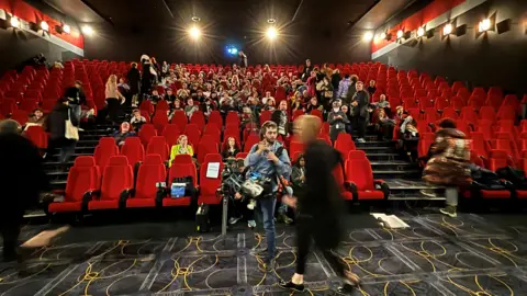 BBC A cinema theatre with red seats and a patterned carpet. People take their seats.