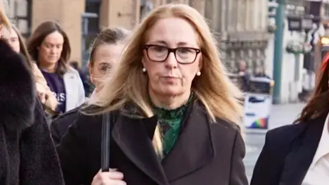 BBC Alison Kelly, who has shoulder length reddish blonde hair and wears dark rimmed glasses and a green top under a black jacket, is pictured as she walks towards Liverpool Town Hall carrying a black handbag on her shoulder and pursing her lips in a serious expression