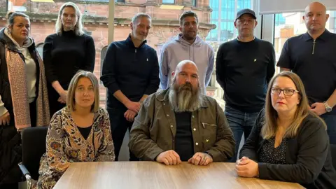 BBC Seven family members gathered in a room with two lawyers. Seated at a table are, from left to right, Louise Slorance, Mark Bisset and Maureen Dynes. Standing behind them are, from left to right, Samantha McDowall; lawyers Stephanie Spencer and Patrick McGuire; Kevin McDowall; Kenneth Murdoch; and David Campbell.