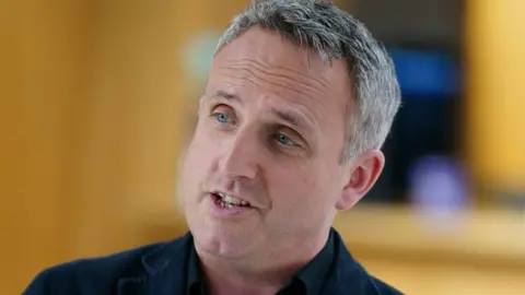 PA Media The leader of the Scottish Liberal democrats, Alex Cole-Hamilton smiles as he look away from the camera. He has grey hair and blue eyes. He appears to be in the Scottish parliaments building but the background is blurred.