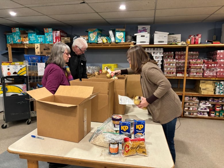 Volunteers sort canned goods and pasta into boxes in a basement food bank with shelves of cans in the background