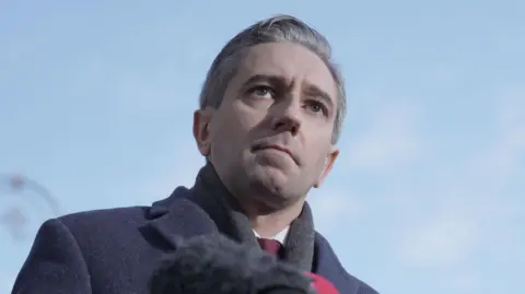 Brian Lawless/PA Wire Simon Harris addressing reporters in Dublin on 20 November.  He is wearing a dark grey coat, a grey scarf, a light shirt and a maroon tie. 