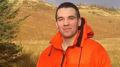 Facebook MacDonald smiles at the camera. He is wearing a bright orange work jacket. He is outside and standing in a rural landscape with a hill doted with trees behind him.