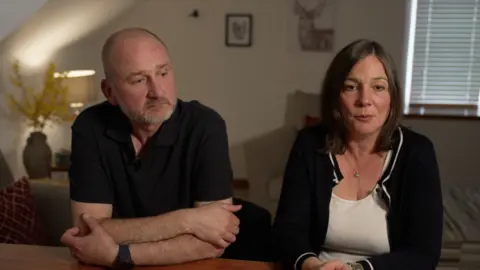 BBC Mark Dowey in a black polo shirt leans on a table looking off to the side in thought, beside his wife Ros in a white top and black cardigan who is looking into the camera. They sit in a light lounge with a lamp on in the background and the blinds drawn.