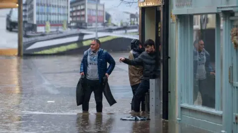 Getty Images Three men stood outside a shop on Mill Street in Pontypridd, wading through flood water. One, wearing a blue coat, is stood in the street. Water can be seend to be entering one property on the right hand side.