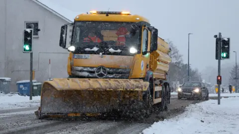 Reuters A yellow snow plough on a snow covered street with a car behind and traffic lights alongside.