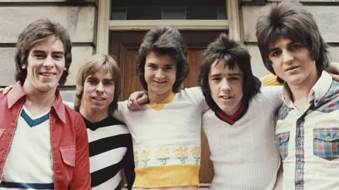 Getty Images The five members of the Bay City Rollers pose arm-in-arm for the camera. Alan wears a trendy mullet with a white sweater and a red jacket with large lapels, Derek, also sporting a mullet, wears a black and white striped jumper, Les, in an impressive bouffant hairstyle, wears a tight white sweater with yellow flower trim, Woody leans forward with his spiky mullet and a white cricket jumper, and Eric keeps it tartan with a white, tartan-trimmed shirt and an impressively layered face-framing hairstyle. They stand outside with a closed wooden door behind them.