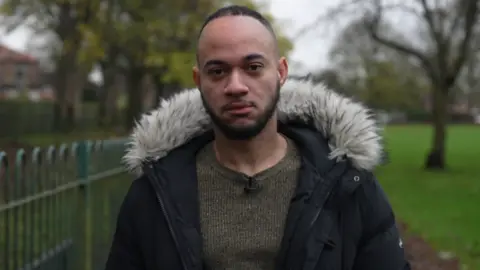 BBC 26-year-old Derai with a serious facial expression. He is wearing a brown woollen jumper and a dark coat with an artificial fur collar.