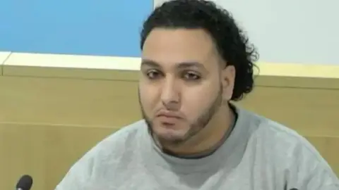 Manchester Arena Inquiry Abdalraouf Abdallah sat in a public inquiry, wearing a grey top and staring straight into the camera. He has curly black hair and a chin strap beard.