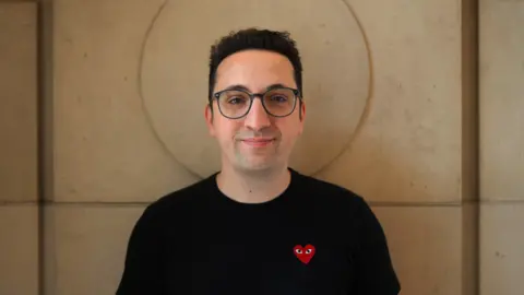 Adam Riches A man with short black hair and glasses and a black t-shirt stands against a sand-coloured wall. His head is perfectly framed by a circular indentation carved into the large stone block.