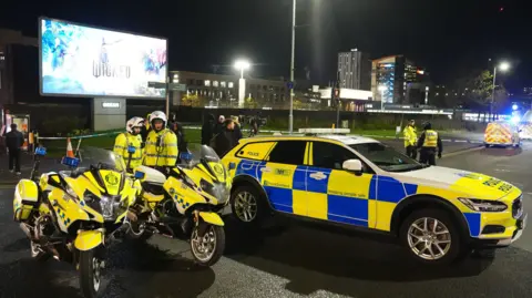 PA Media Police bikes and a car blocking the road close to the bus station with flats in the background. It is nighttime. Officers stand around wearing hi vis.