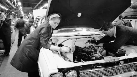 Getty Images A black and white photo of workers helping to build a Vauxhall Victor. A woman wearing a headband and technician's coat is working on a car engine.