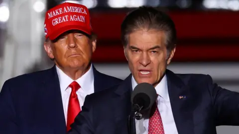 Reuters Mehmet Oz speaking into a microphone and gesturing as Donald Trump behind him looks on, wearing a red "Make America Great Again" hat. 