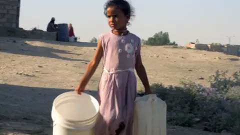 BBC A young girl wearing a pink dress carries three empty plastic buckets and a cannister as she walks outdoors in Hassakeh