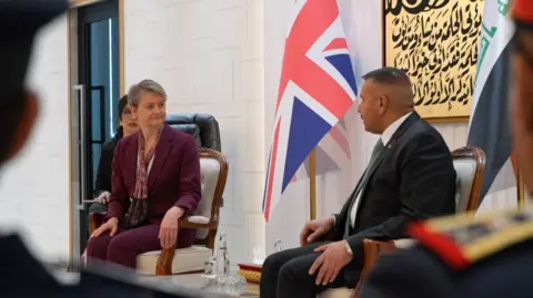 Lee Durant/BBC Yvette Cooper and Abdul Amir Al Shammari sitting in chairs side-by-side in front of the UK and Iraqi flags. 