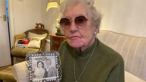 BBC Win Crew holding a photo of her and Reginald when they were younger. She is wearing glasses, a dark green jumper, and necklace, and is sitting on a sofa.